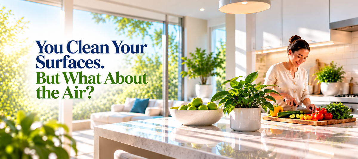 A woman in a bright modern kitchen prepares food at the counter, with large windows showing greenery outside and overlaid text reading, 'You Clean Your Surfaces. But What About the Air?'— Now Treat Both at Once